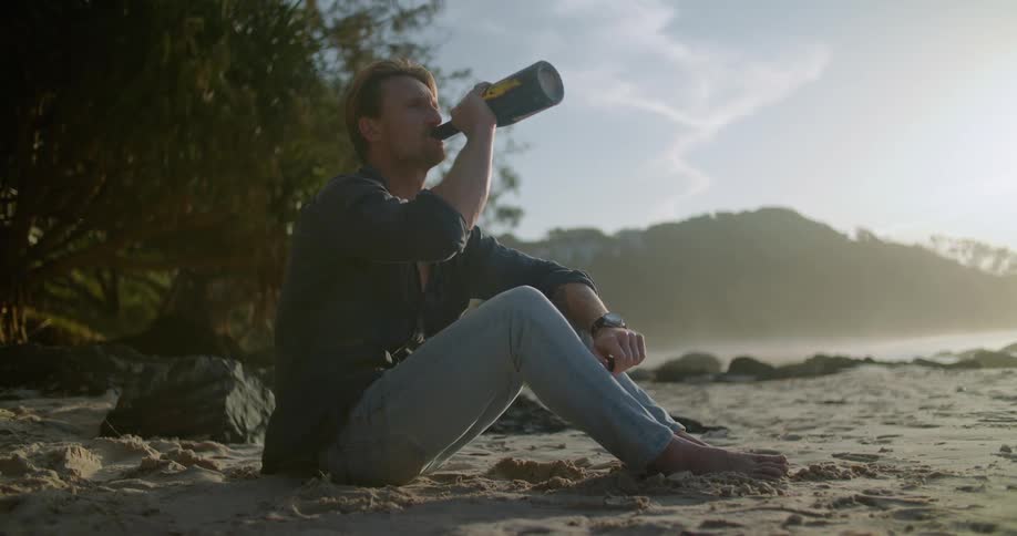 A man is sitting at the beach and drinking an alcoholic beverage