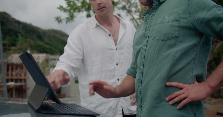 A man leans forward pointing to a tablet while talking with another man