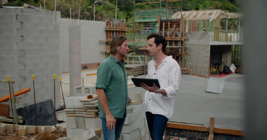 Two men standing at a construction site talking and gesturing while one holds a tablet