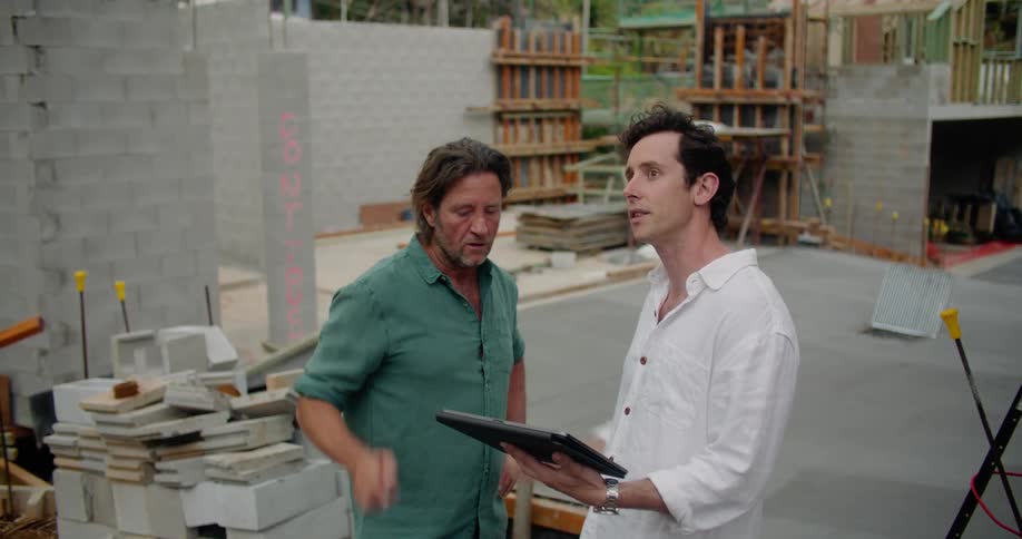Two men stand together at construction site holding discussion while consulting tablet