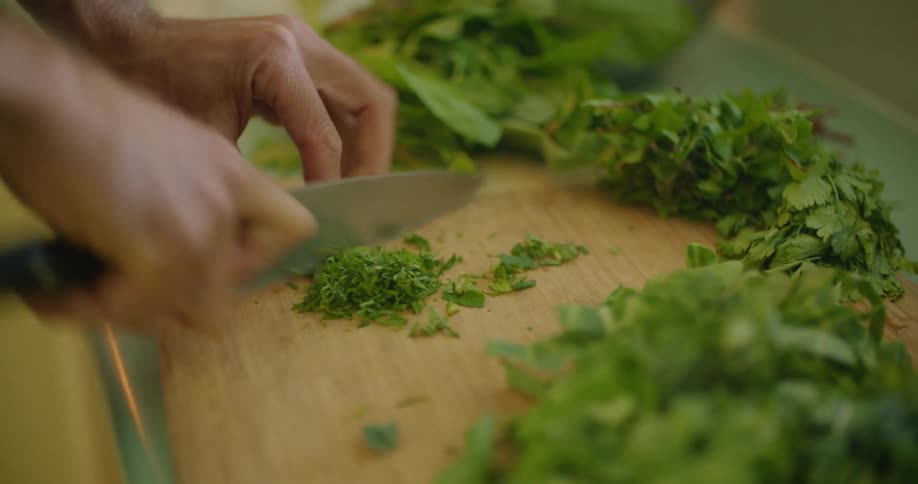 Man chopping herbs finely over a wooden board with a sharp kitchen knife