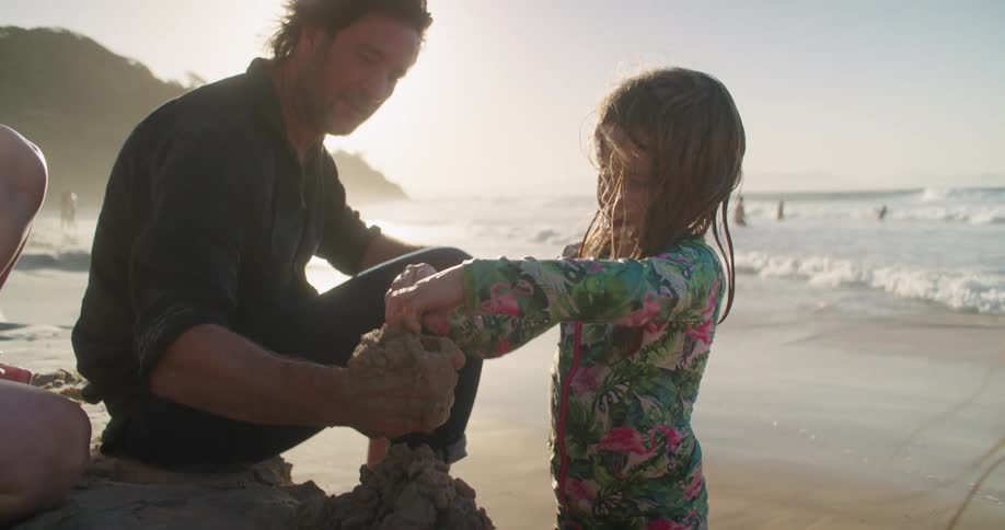A dad sitting on the beach as his daughter piles wet sand into his hand