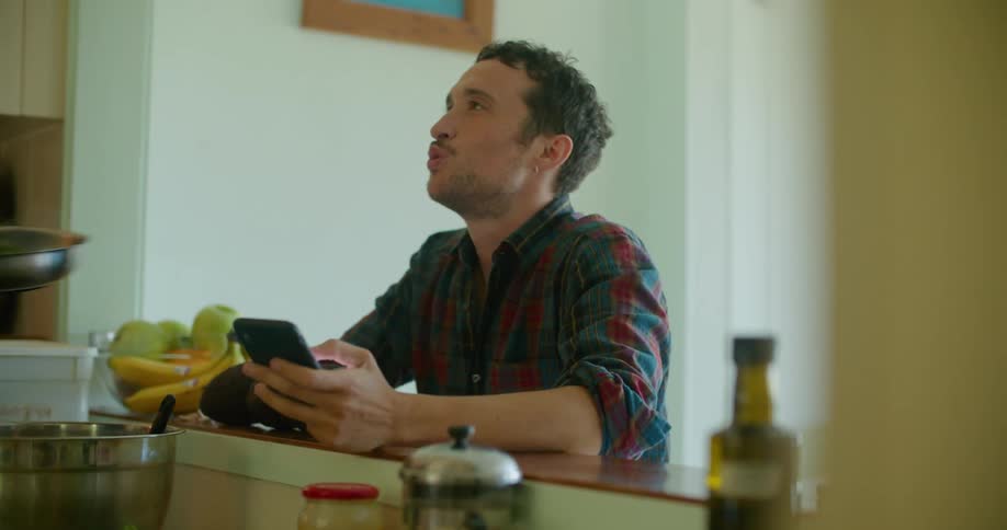 A man is sitting at a kitchen bar looking at his cellphone and talking when someone gives him a skillet of food to sniff