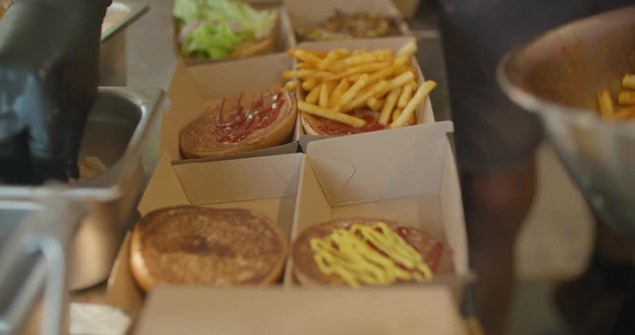 A person puts fries in cardboard containers as another person garnishes buns in a fast food restaurant