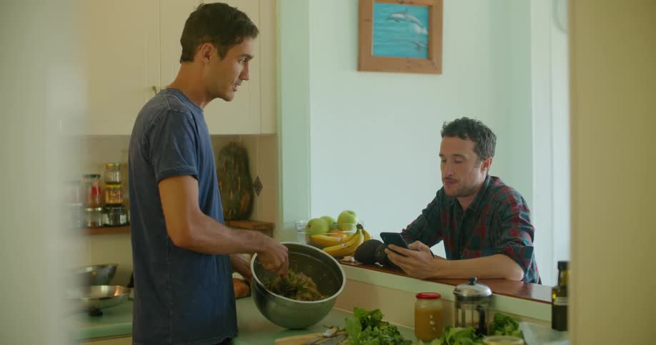 A man shows another man his cell phone while he is cooking