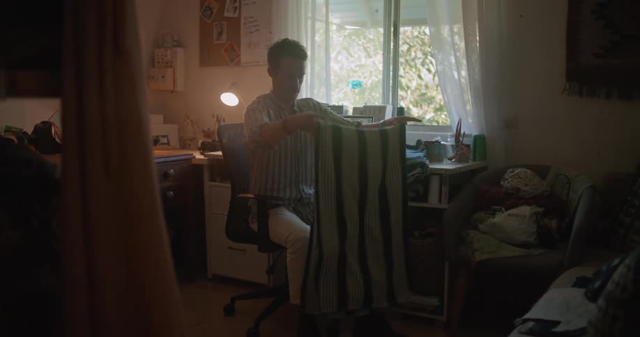 Man folds cloth while sitting at table with sewing machine and computer