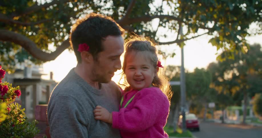 A father is standing outside holding his daughter and they give each other kisses on the cheek