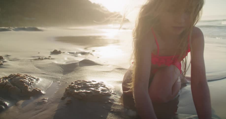 Girl wearing bikini kneels on beach playing with sand as parent stands up nearby