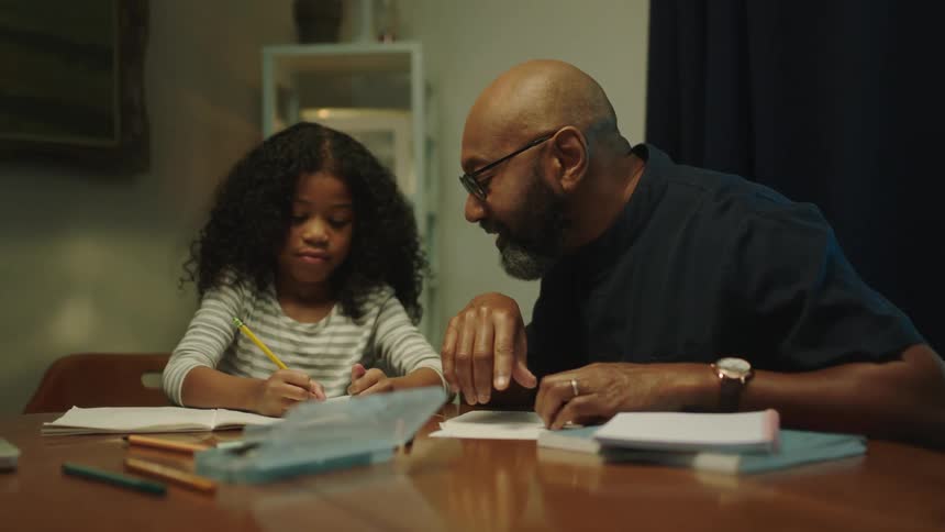 A father helps his daughter do homework at the dining room table covered in books and pencils