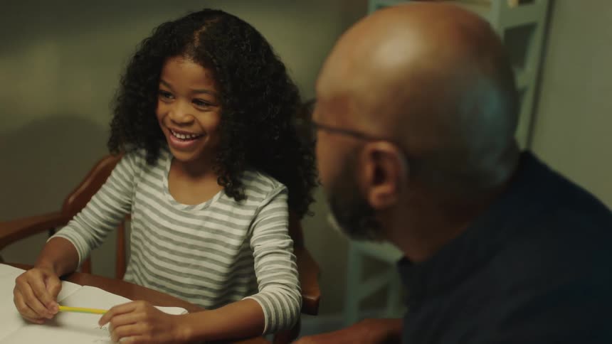 A girl smiles as her dad talks then she writes in her notebook