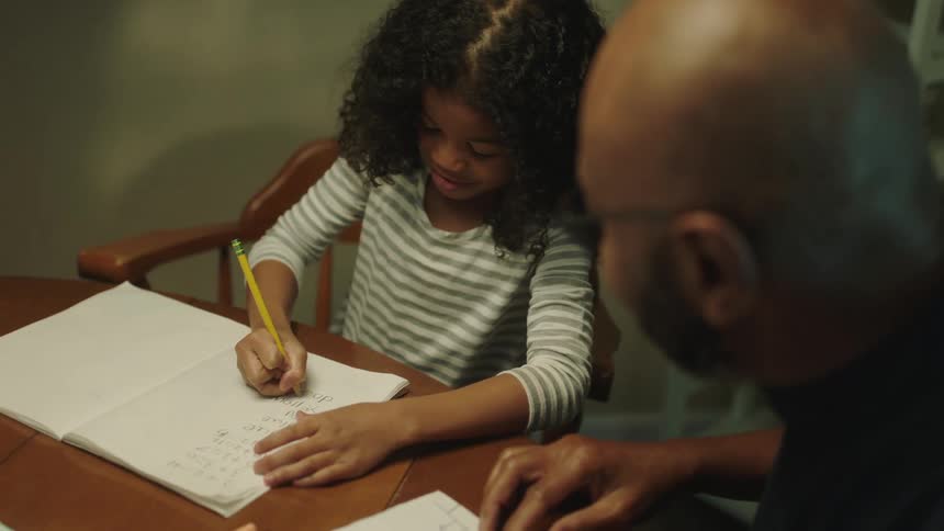 A father is watching his daughter do her homework at a table in their home at night