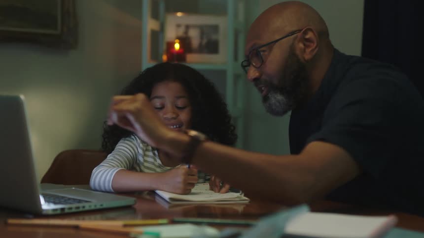 A man is pointing at a laptop while helping his daughter with homework