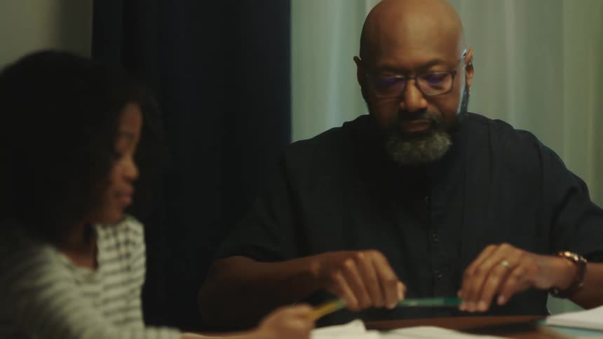 Father helps daughter with homework while sitting at a table in the living room
