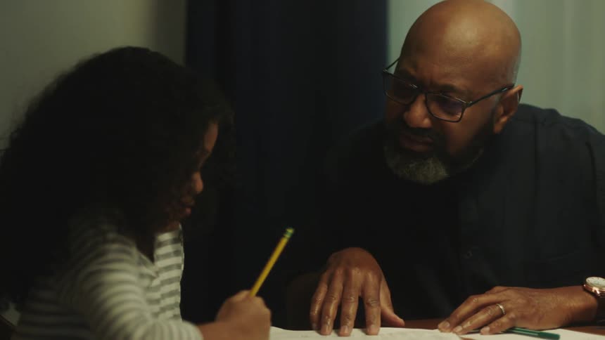 Bald teacher with glasses beard and black shirt teaches sits at table teaching girl with curly hair