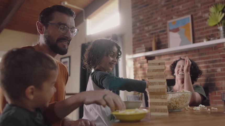 Family sits at table with wooden block stacking puzzle laughing and throwing popcorn
