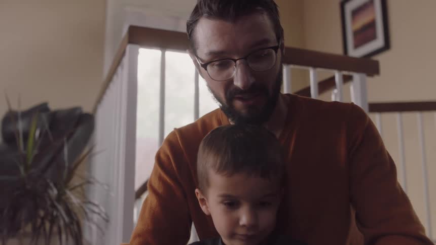 A father and son are reading a book together in their home while sitting on the floor in the daytime