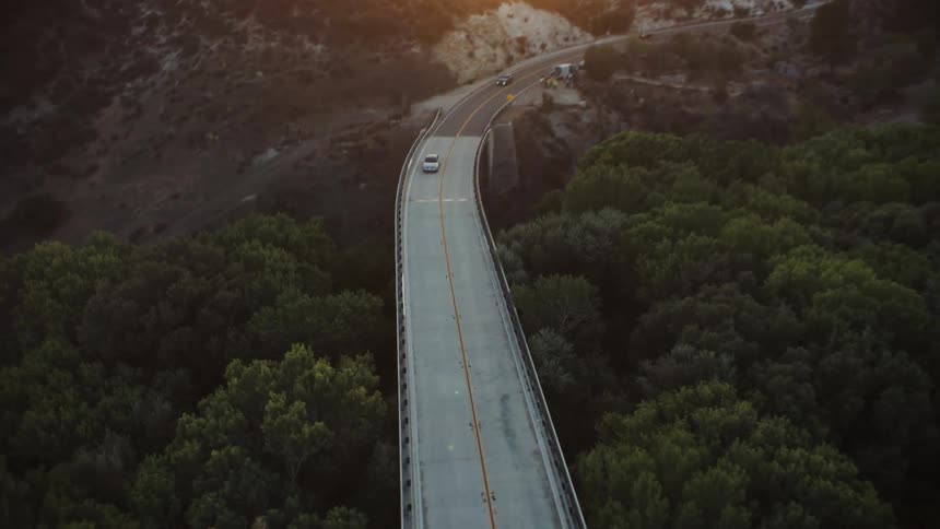The sun shines over a mountain as a silver car is traveling on a bridge with trees below