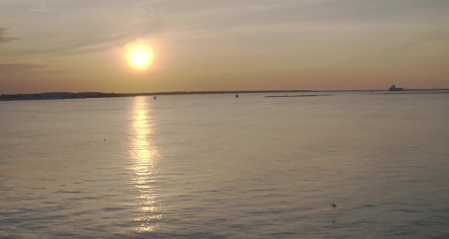 Seagulls are flying over the water as the sun is setting in the distance