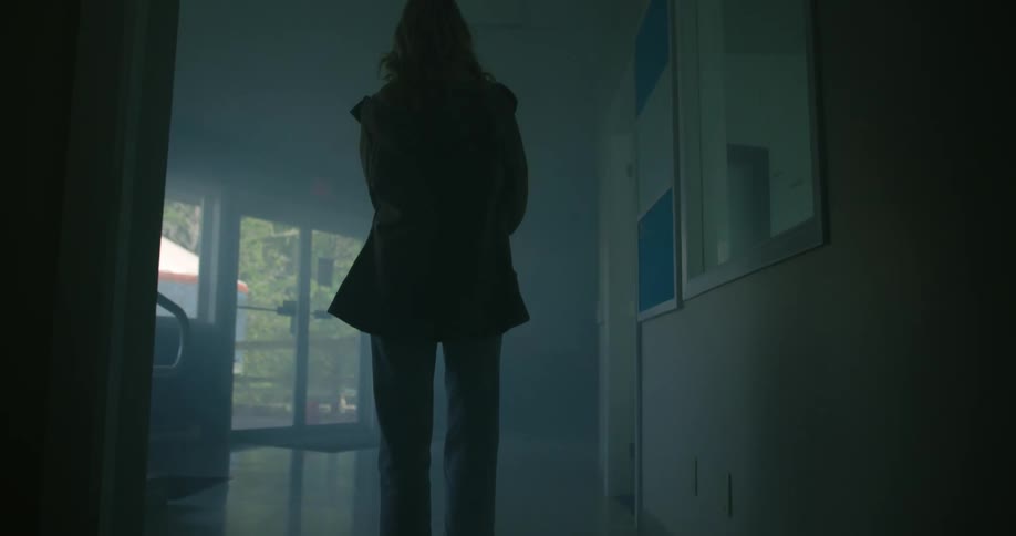 The back of a woman walking from a darkened hallway toward glass exit doors with sunlight shining