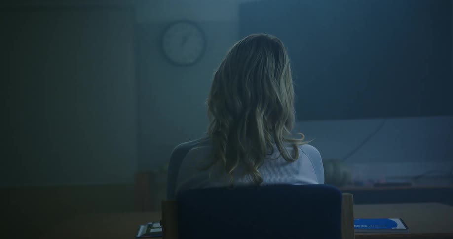 A blond hared female person is sitting in a chair at a table facing a wall with a clock and holding a pen in her right hand