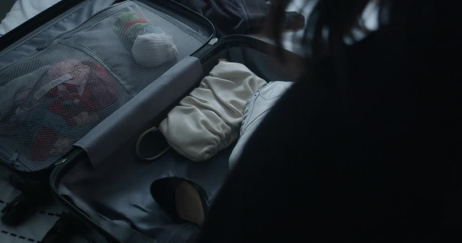A woman at home packs shoes and purses in to a suitcase on a bed