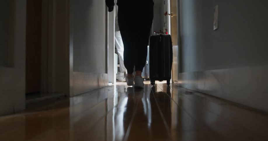 Two people walk down a hallway of a house towards a kitchen rolling their suitcases