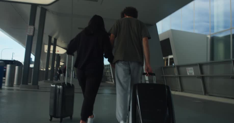 A man and woman pull their suitcases behind them as they walk towards the airport and the man reaches over and puts his arm around the woman and then holds her hand
