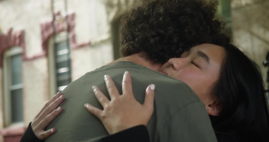 A woman smiles as she leans in for a hug from a man in front of a building
