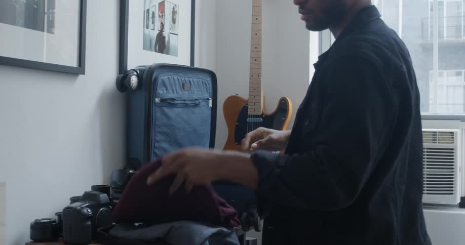 Man folds clothes into wheeled suitcase sitting on table near guitar