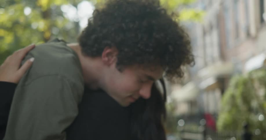 Man with curly hair and green t shirt and woman with straight hair and black sweater cuddle and kiss each other in the street next to apartment buildings