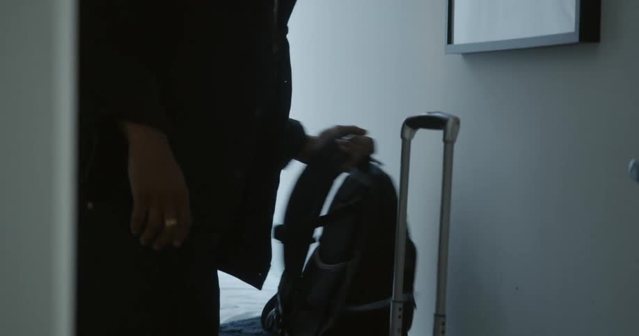 A man picks up his backpack and grabs his suitcase handle walking out of a bedroom