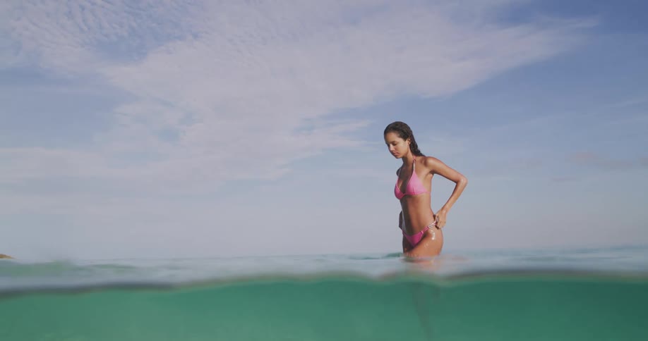 A beautiful woman in a pink bikini walks through clear water toward the beach