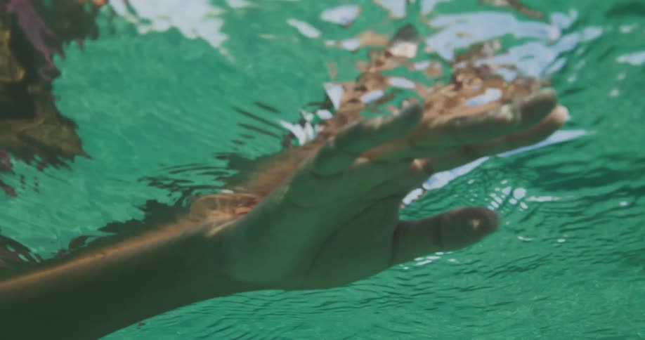 A persons arm and hand is moving in the water causing ripples as the sunlight shines from above