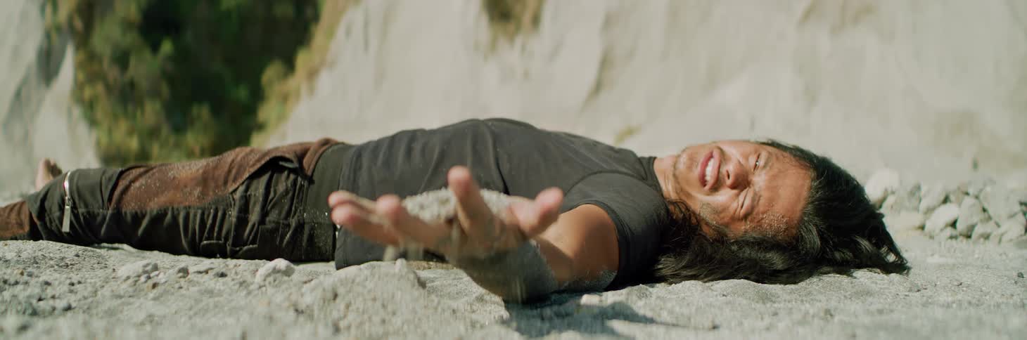 A man lies in sand at the bottom of a canyon and grimaces in pain as he grabs a handful of sand and squeezes it and lets it fall through his fingers