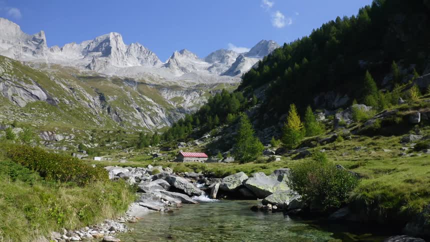 River flows through grassy valley past red roofed building nestled against forest covered mountain
