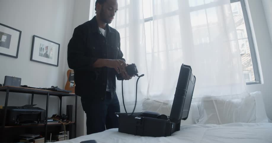 Male photographer packs camera into case in bedroom with guitar on wall next to table