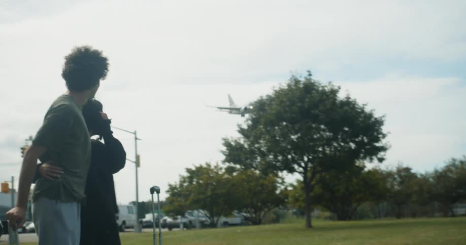 Two people are standing near a road arm in arm and watch a plane come in for a landing