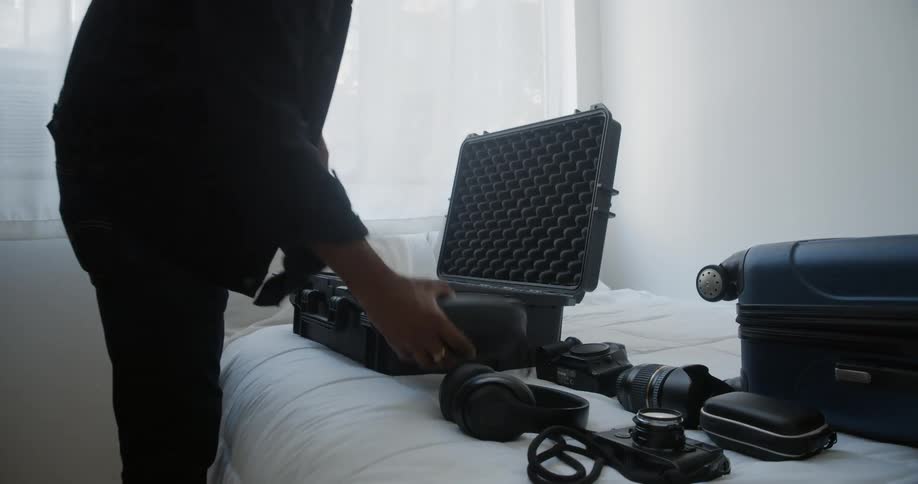 Man packs camera and other items in luggage sitting on bed