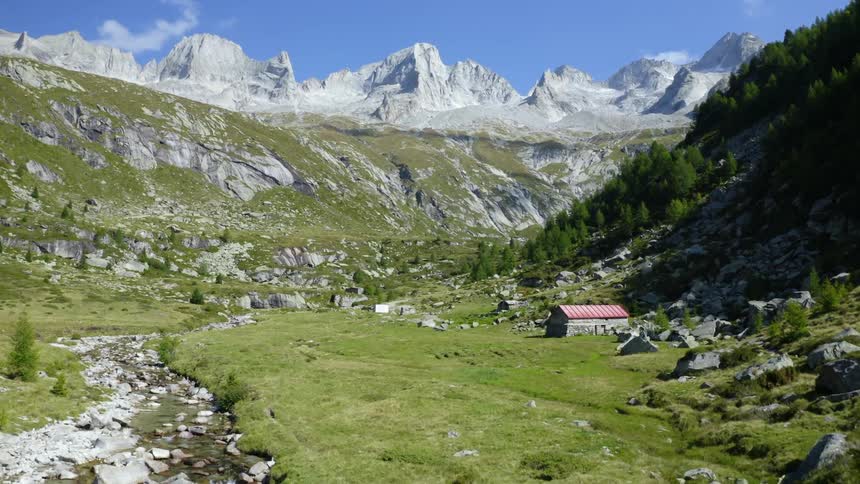 A mountain streams flows from green slopes near a farm building under a bright blue sky