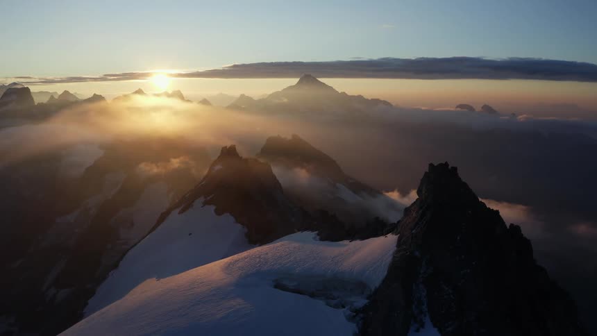 The sun is shining brightly on snow covered mountains with peaks high in a cloudy sky