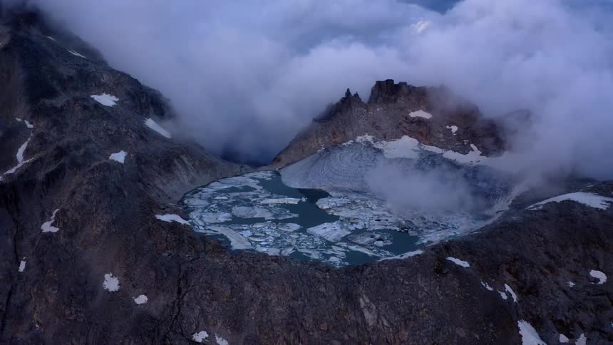 An aerial view of a pool of water and ice on a mountain with low clouds around it