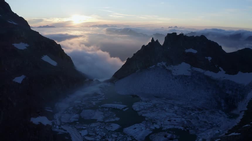 An aerial view of a pool of water and ice on an mountain landscape with clouds below and the sun rising early in the morning
