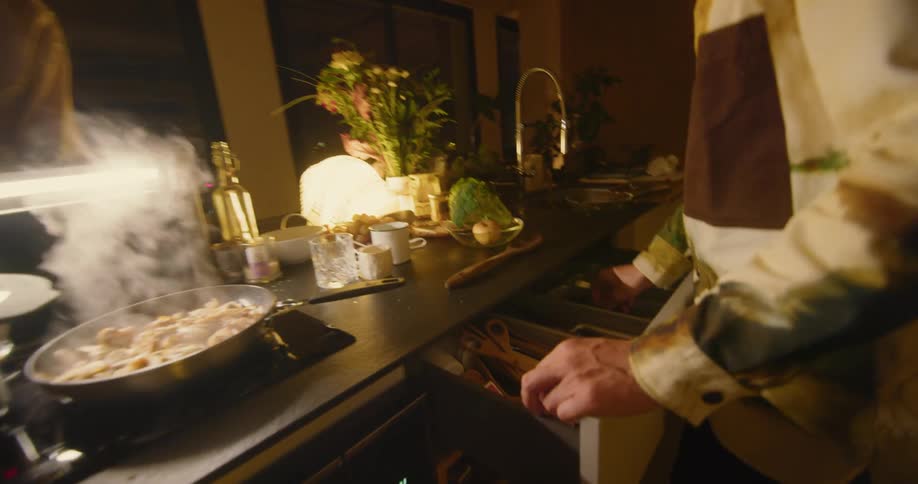 A man is preparing food in an apartment at night with a woman sitting nearby at the counter