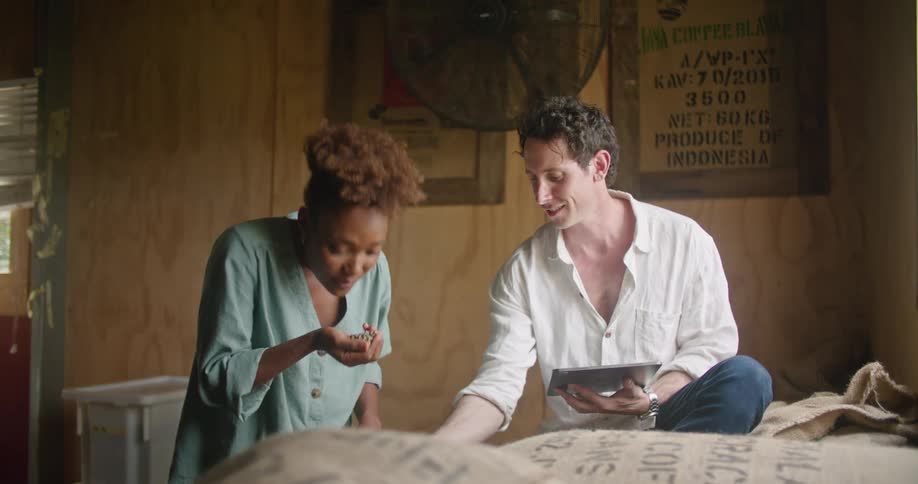 Female worker with curly hair and and turquoise shirt smells coffee beans and talks to man with white t shirt and jeans using tablet in coffee warehouse