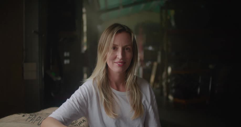 Blonde woman with white t shirt leans against coffee sacks looking at camera smiling in coffee warehouse