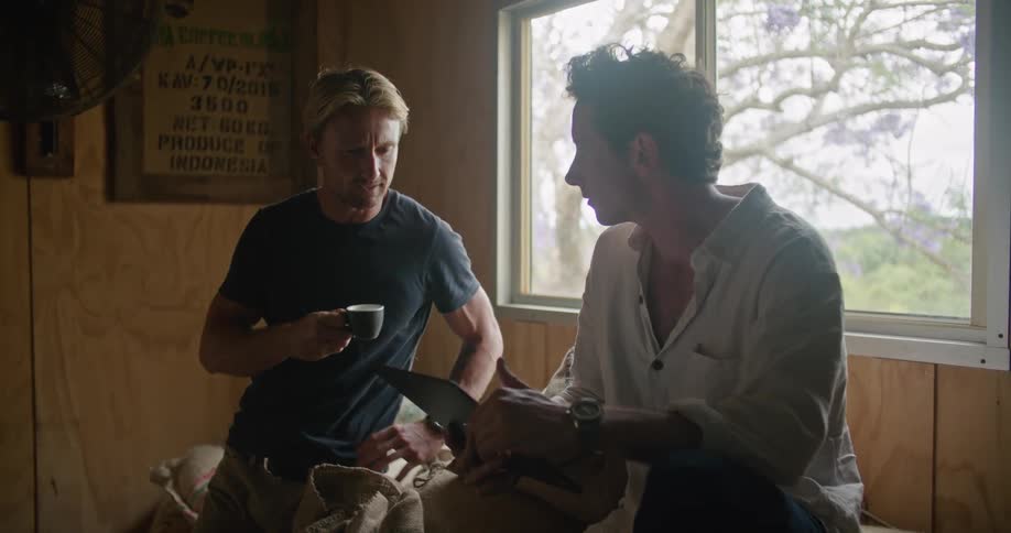 A man drinks coffee while chatting with a colleague holding a tablet