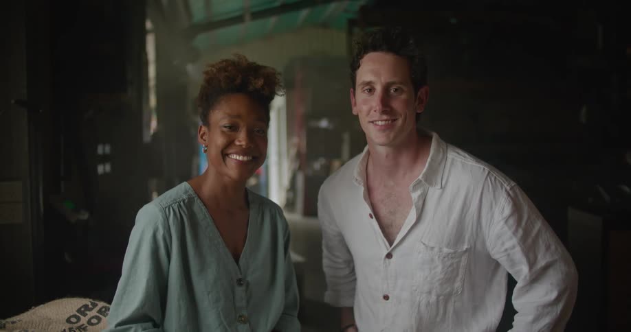 Man with white shirt and woman with curly hair earring and light blue shirt face the camera with eye contact smiling next to coffee bags in storage facility