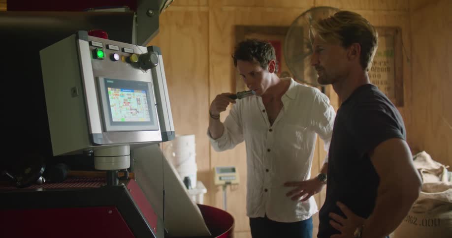 A man removes a sample from the industrial roaster to test the aroma as he discusses business with a coworker