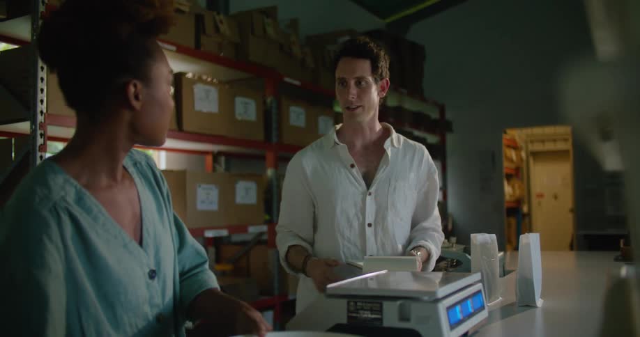 Female worker with curly hair and and turquoise shirt works while man with white shirt checking notebook in coffee warehouse