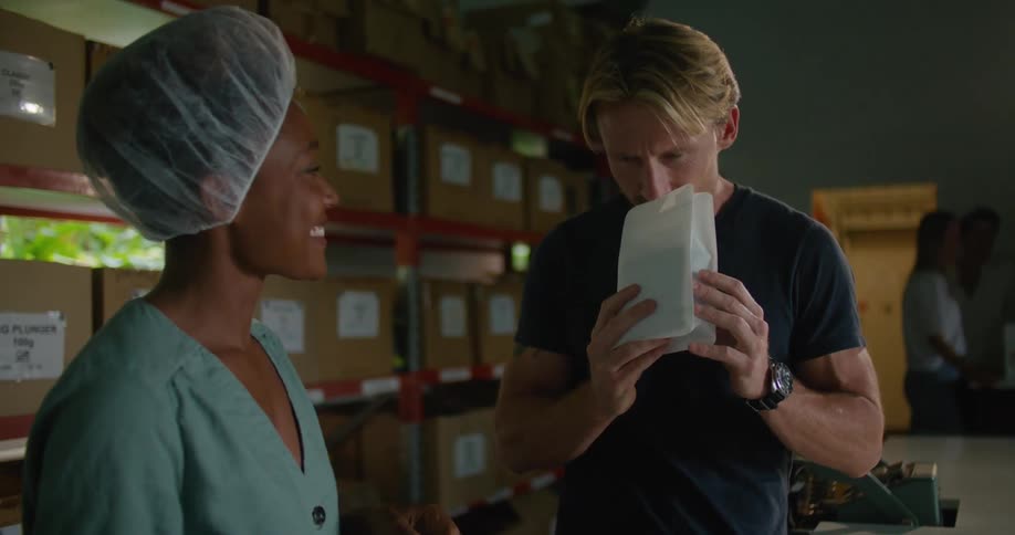 Female worker with hair bonnet and and turquoise shirt gives coffee beans to blonde man with dark blue t shirt to smell in coffee warehouse